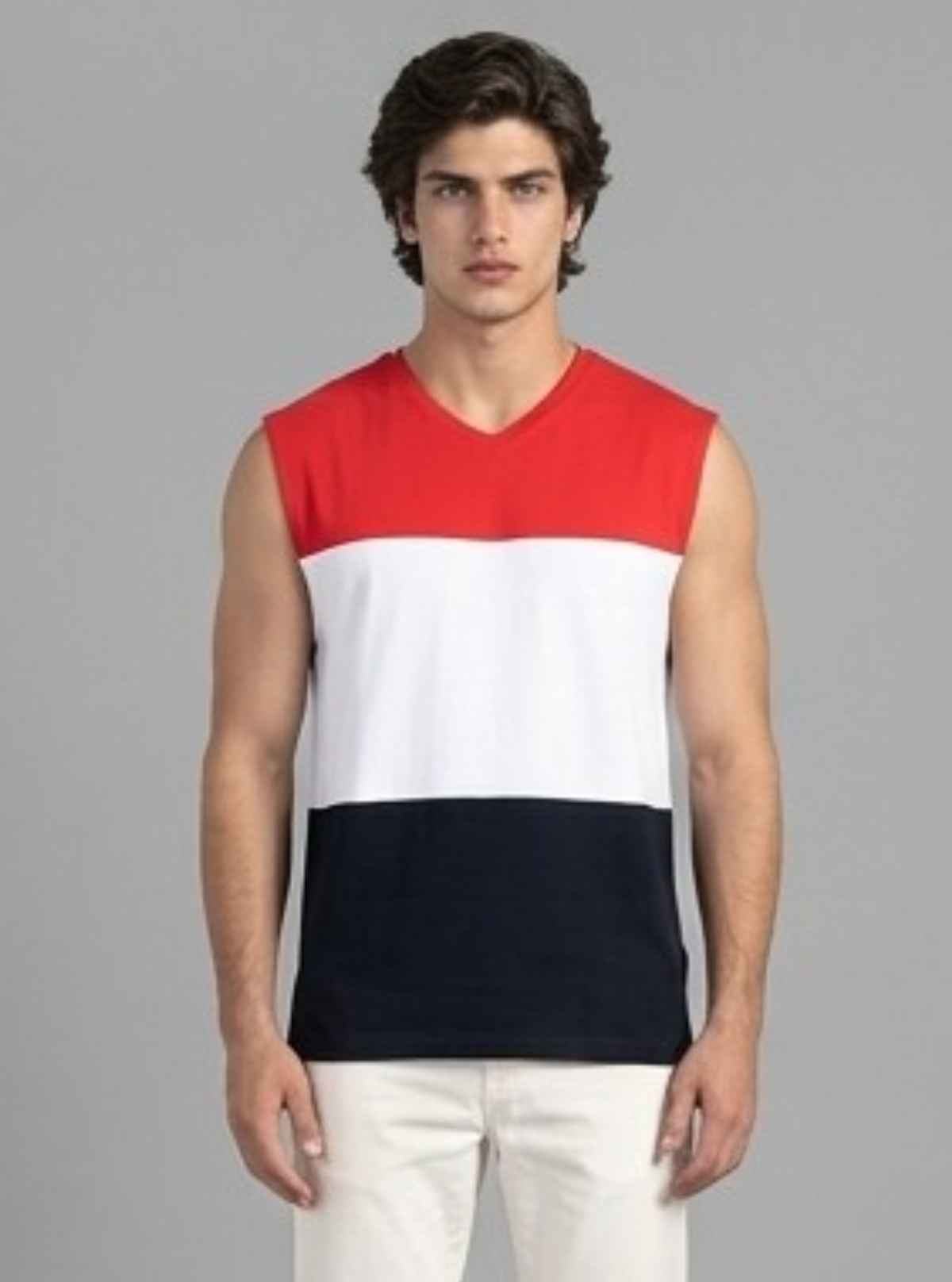 A man modeling a sleeveless V-neck knit top with horizontal red, white, and navy color-block panels, paired with off-white pants, against a solid gray background.